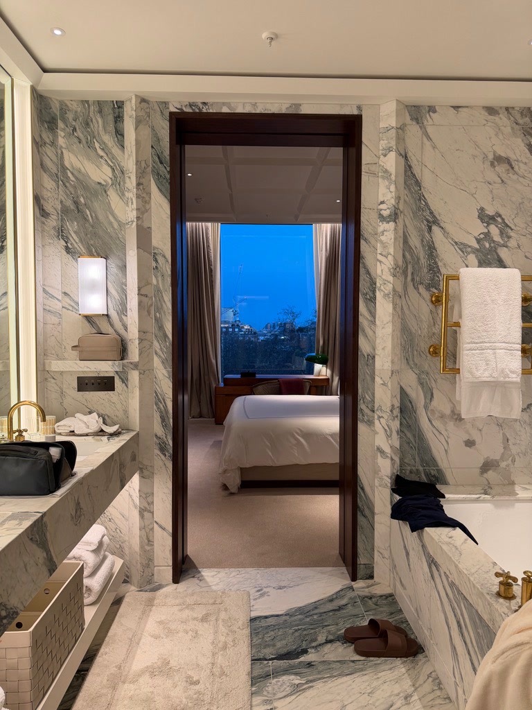 A marble bathroom at dusk, looking through a dark wood doorway to a made bed and a tall window over the city